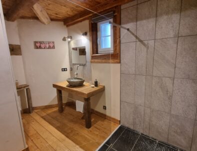 At St Maurice, in the Rimandoule room, you will particularly appreciate this large walk-in shower in a warm wooden decor with exposed beams. A gray stone washbasin is placed on an old reclaimed wooden support.